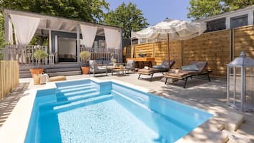 Piscina con sillones de descanso y sombrillas, rodeada de cubierta de madera y una zona de estar techada.
