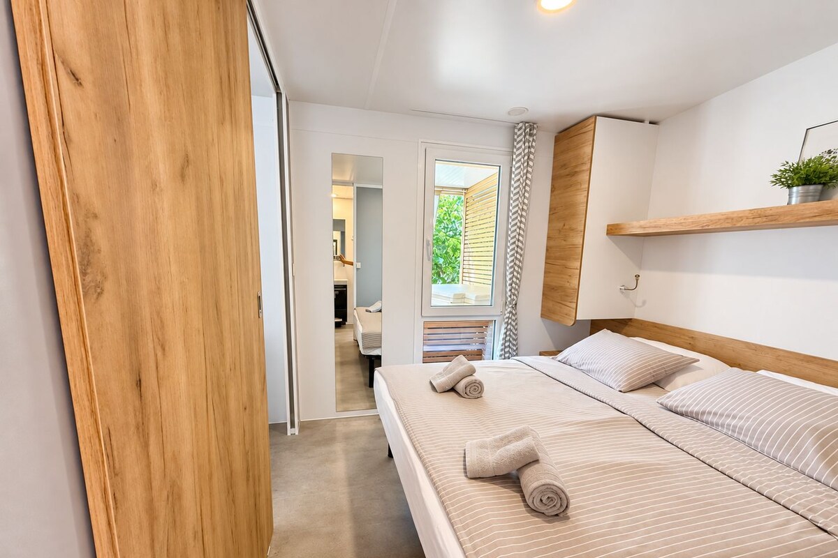 A bedroom with a bed, a wooden wardrobe, a mirror, and a window with a view of greenery outside.