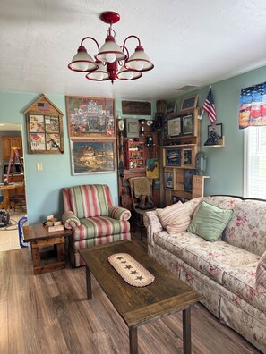 Ein gemütliches Wohnzimmer mit einem gestreiften Sessel, einem blumigen Sofa, einem Holztisch und verschiedenen Wanddekorationen, darunter ein altes Telefon und gerahmte Bilder.