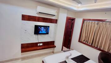 Habitación de hotel con cama, televisor montado en la pared, aire acondicionado, ventilador de techo y ventana con cortinas.