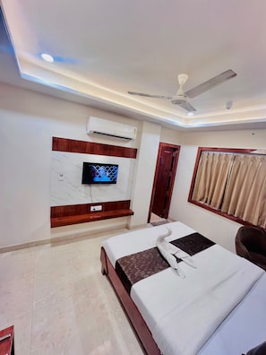 A hotel room with a bed, a television mounted on the wall, an air conditioning unit, a ceiling fan, and a window with curtains. - HOTEL CROSSLANE (KOTA)