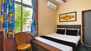 A hotel room with a bed, a chair, a window with curtains, an air conditioning unit, and a painting on the wall.