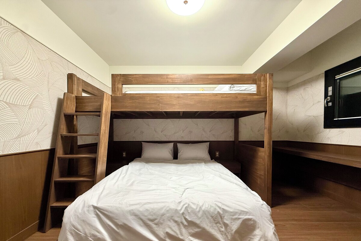 Uma cama de solteiro com escada de madeira, uma cama baixa embaixo e uma televisão na parede.