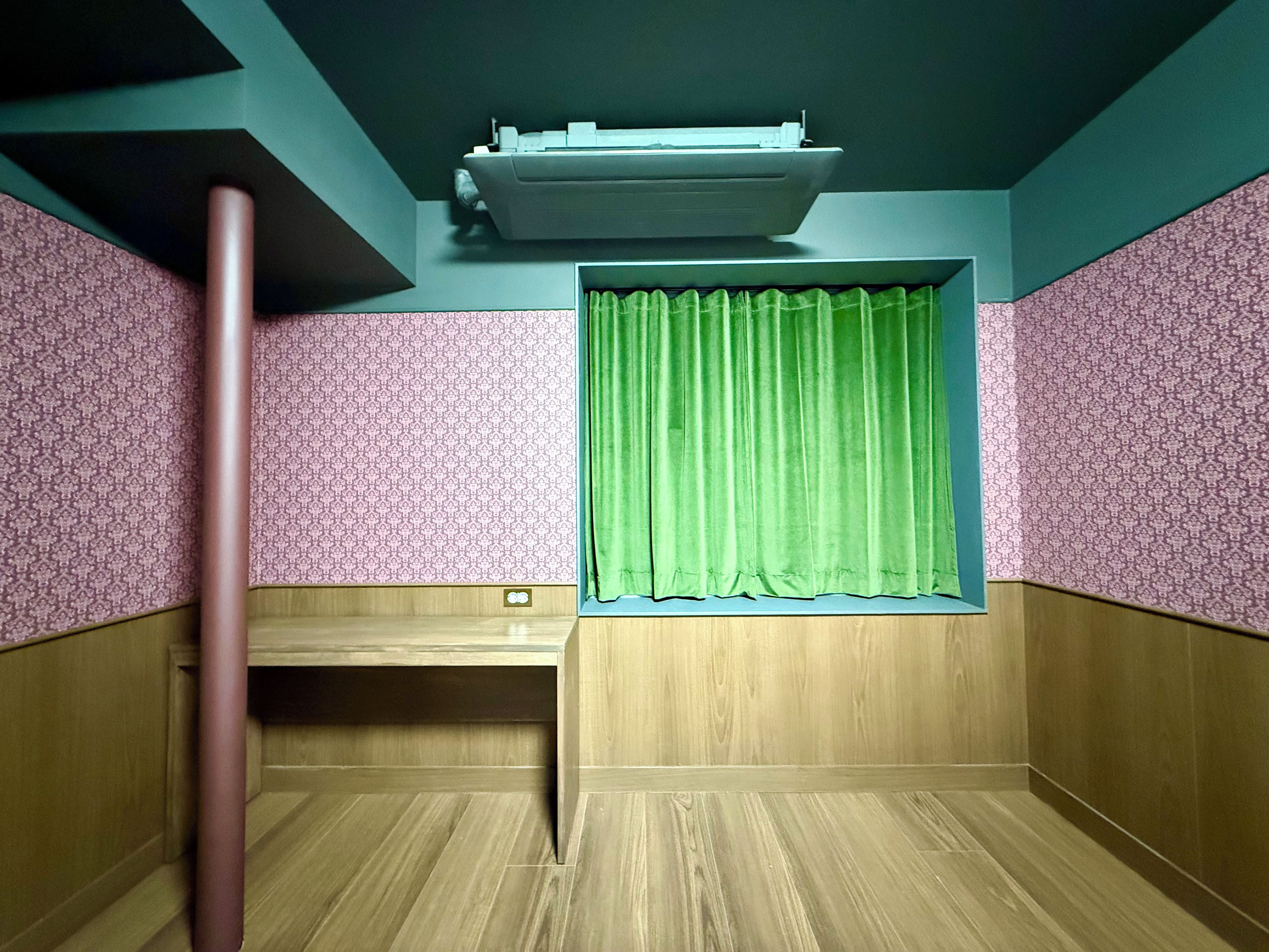 Een kamer met een houten bureau, een groen gordijn, behang met een patroon en een airconditioner aan het plafond.
