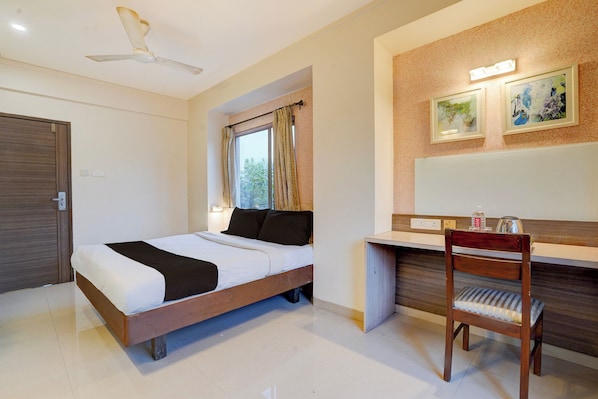Una habitación de hotel con una cama, un escritorio, una silla, un ventilador de techo y dos cuadros en la pared.