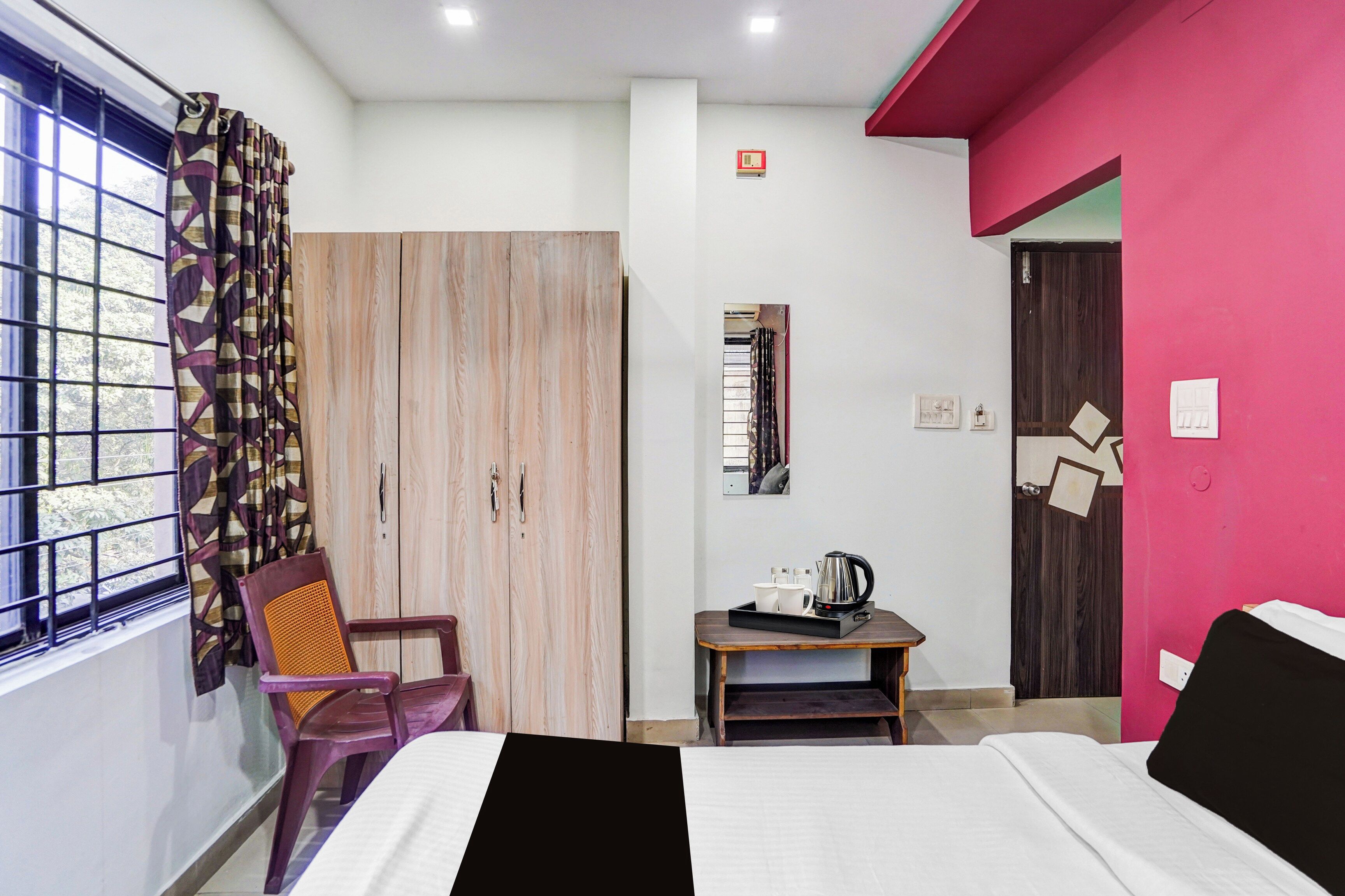 Un dormitorio con cama, una silla, armario, una mesita con hervidor y ventana con cortinas.