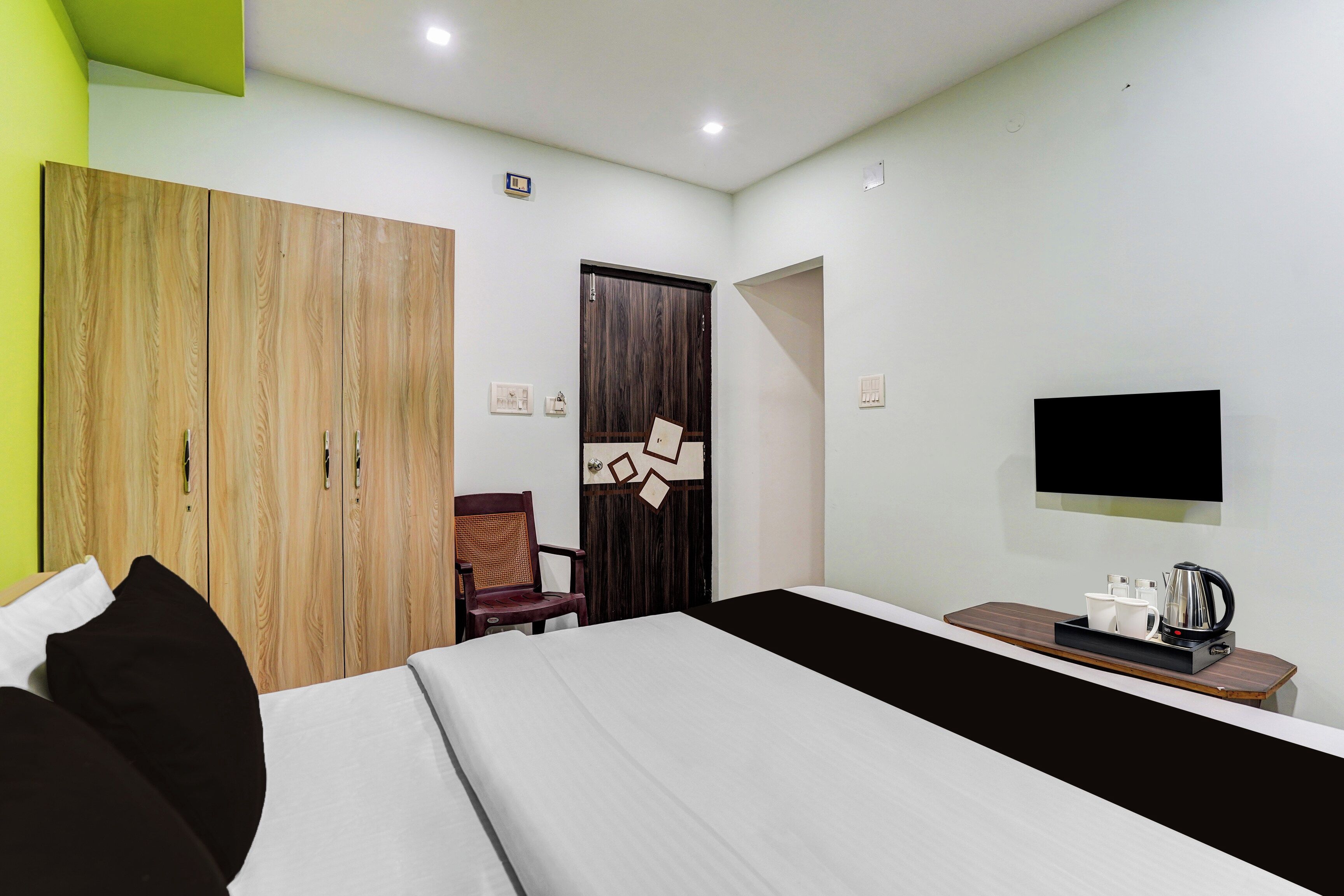 A hotel room with a bed, a wooden wardrobe, a chair, a desk with a kettle, and a flat-screen TV.