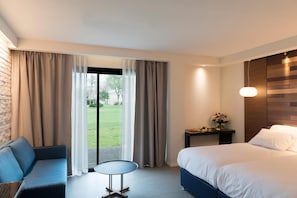 Habitación de hotel con cama, sofá, una mesita y vista a un jardín por la ventana.
