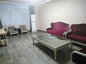 Uma sala de estar com uma mesa de centro de vidro, duas poltronas, um sofá, uma mesinha e uma geladeira.