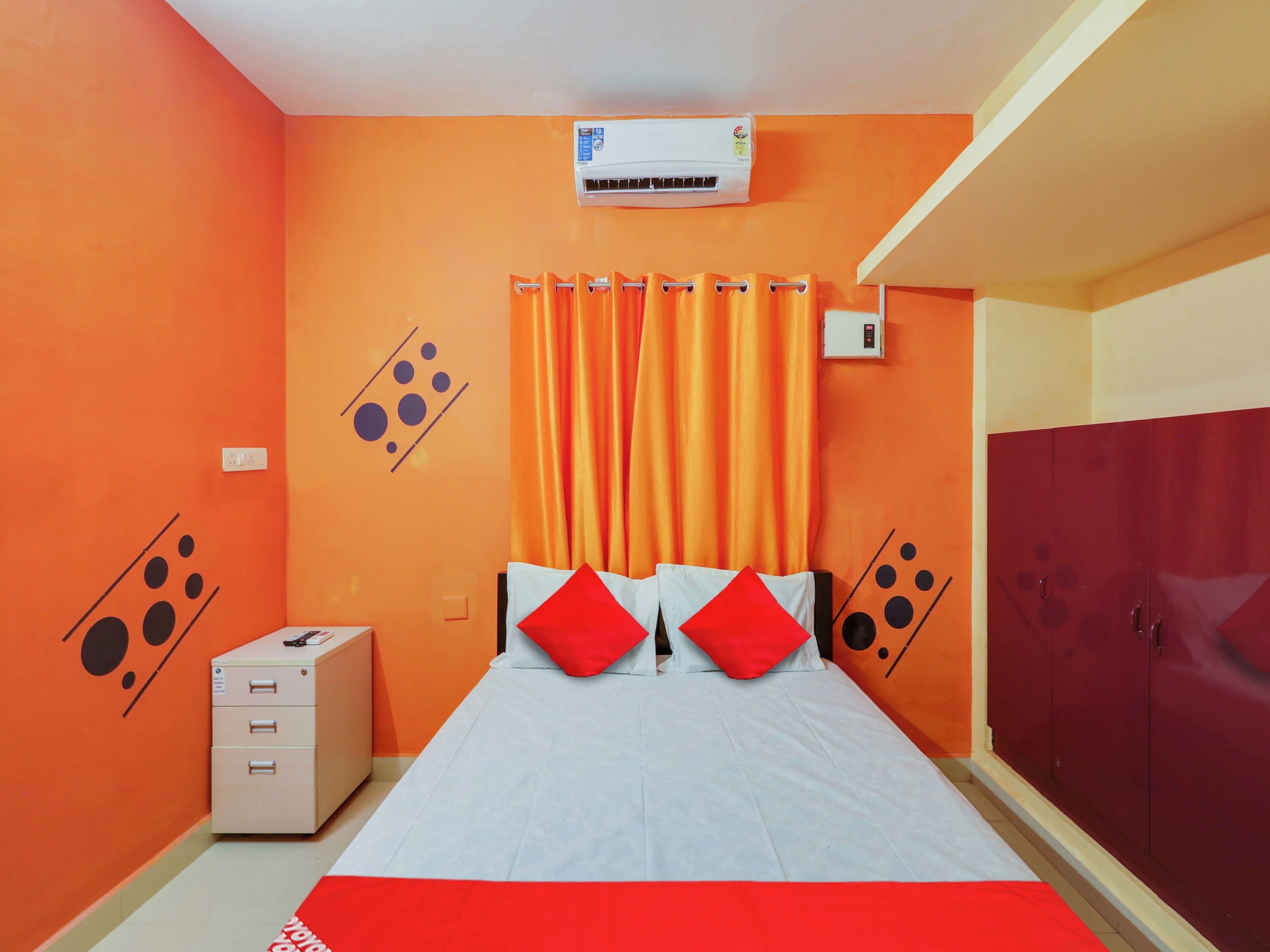 Un dormitorio con pared naranja, cama con ropa de cama blanca y almohadas rojas, un equipo de aire acondicionado, un armario y un escritorio pequeño con cajones.