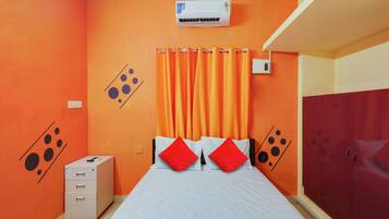 Un dormitorio con pared naranja, cama con ropa de cama blanca y almohadas rojas, un equipo de aire acondicionado, un armario y un escritorio pequeño con cajones.