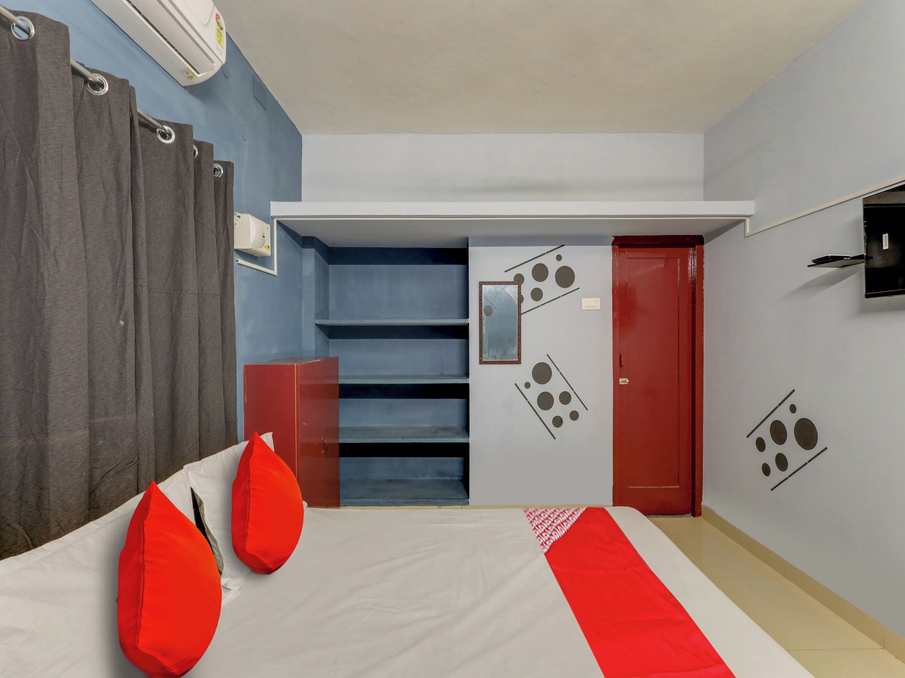 Une chambre d’hôtel avec un lit, une literie rouge et blanche, une armoire rouge, un miroir et un mur orné de motifs abstraits.
