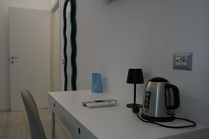 A small, neatly arranged hotel room with a white desk, a kettle, a remote control, and a lamp. - New Listing (Roma)