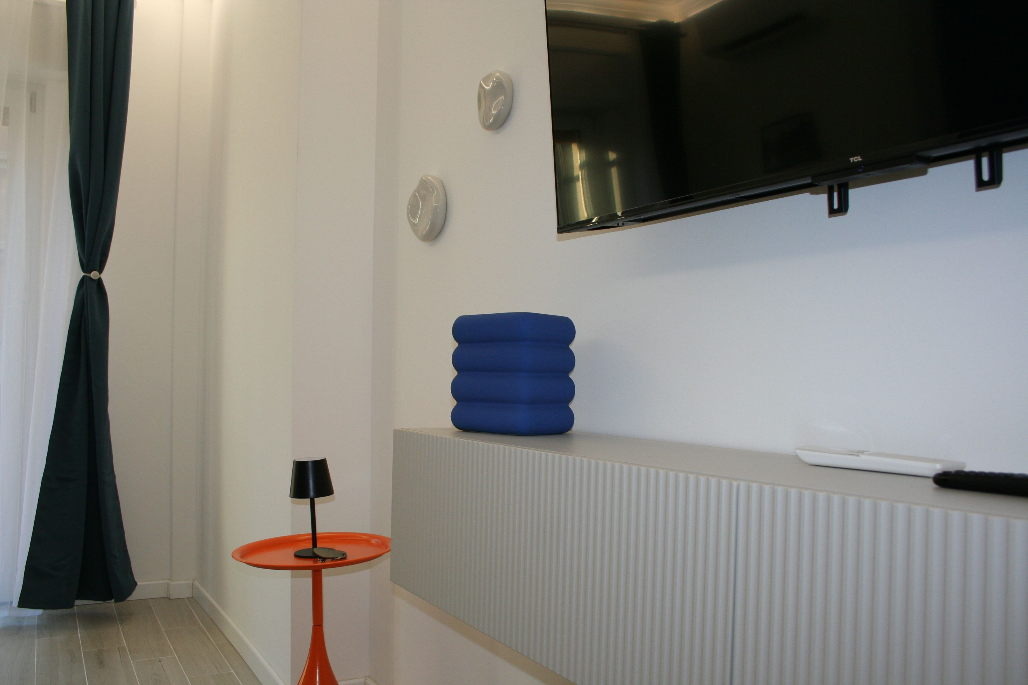 A modern hotel room with a flat-screen TV mounted on the wall, a small orange table with a lamp, and a blue cushion on a white console.