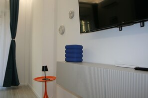 A modern hotel room with a flat-screen TV mounted on the wall, a small orange table with a lamp, and a blue cushion on a white console. - New Listing (Roma)