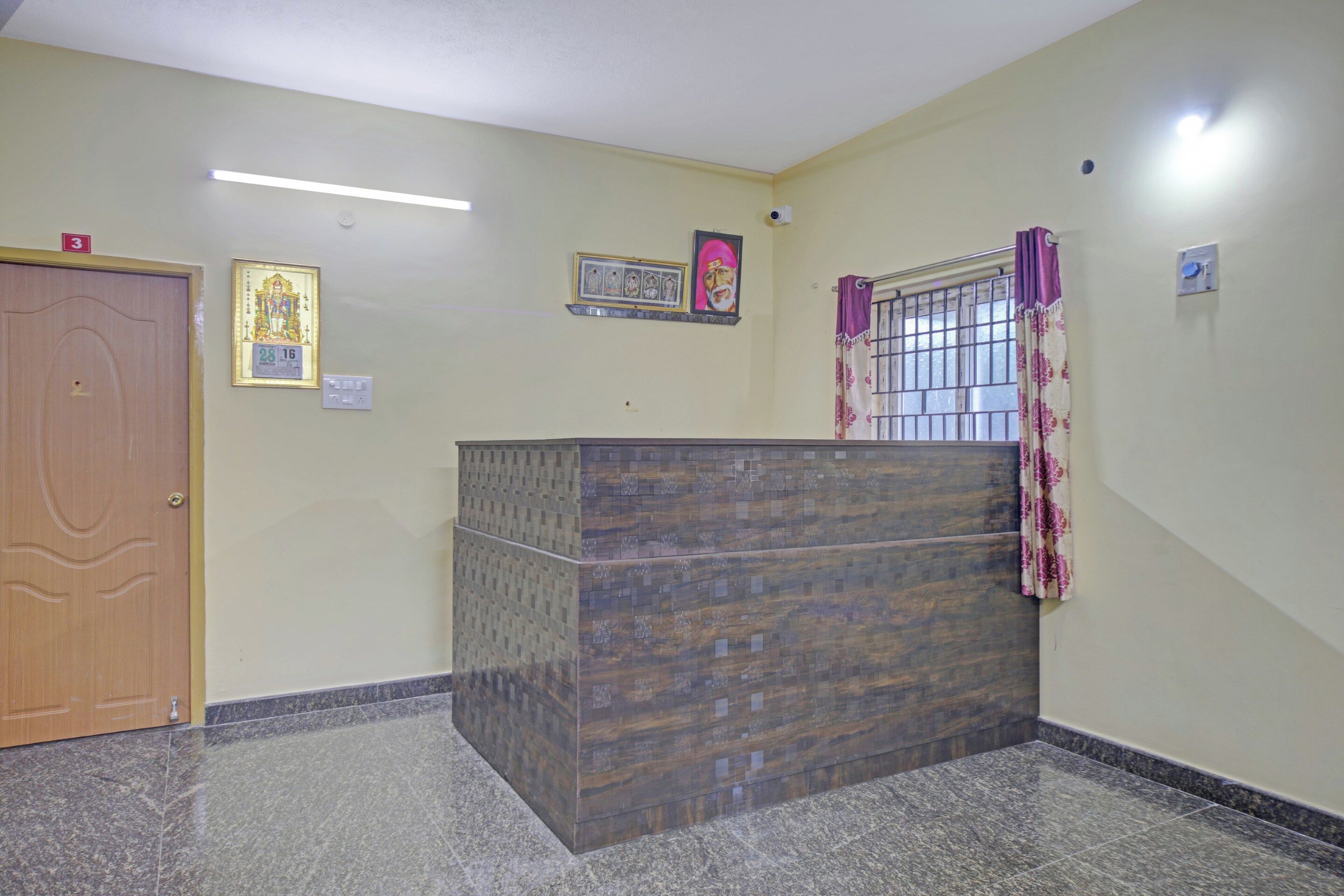 Quarto com porta de madeira, balcão de recepção, luminária fixada na parede e janela com grades e cortinas.