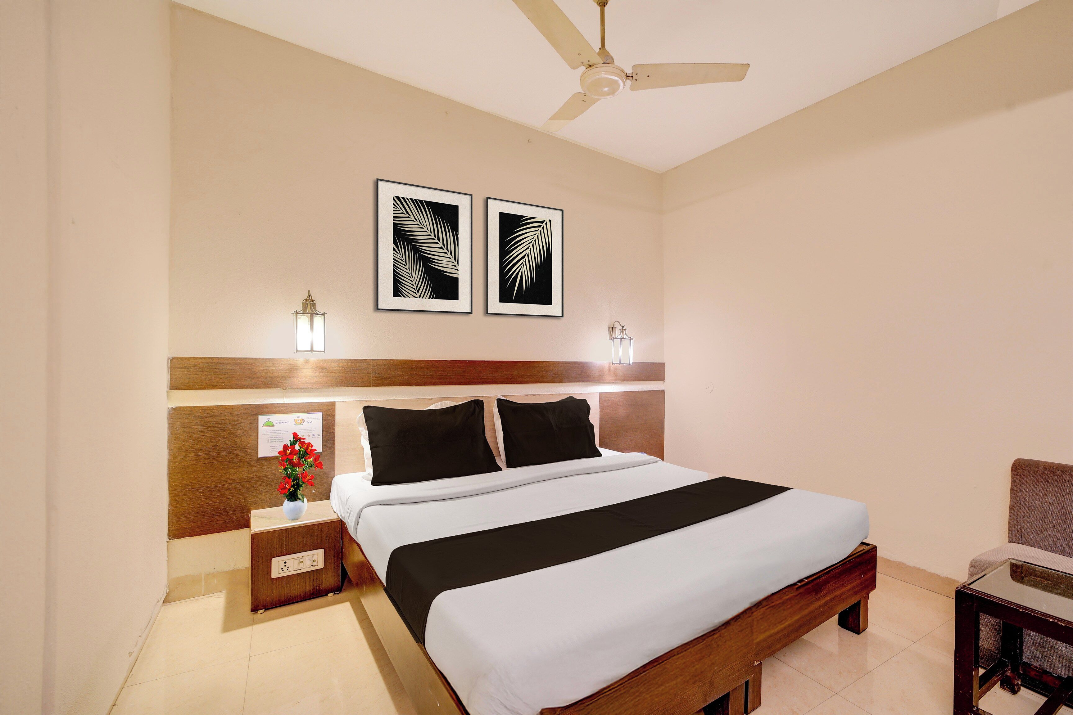 Habitación de hotel con cama, mesita de noche, florero con flores, ventilador de techo y dos cuadros en la pared.