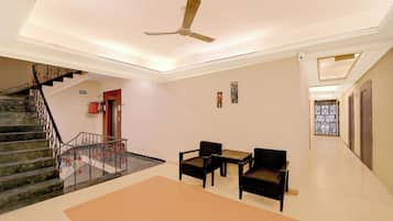 Un hall d’hôtel avec un escalier, un ventilateur de plafond, un panneau indiquant la sortie de secours, et un espace détente comprenant deux fauteuils et une table.