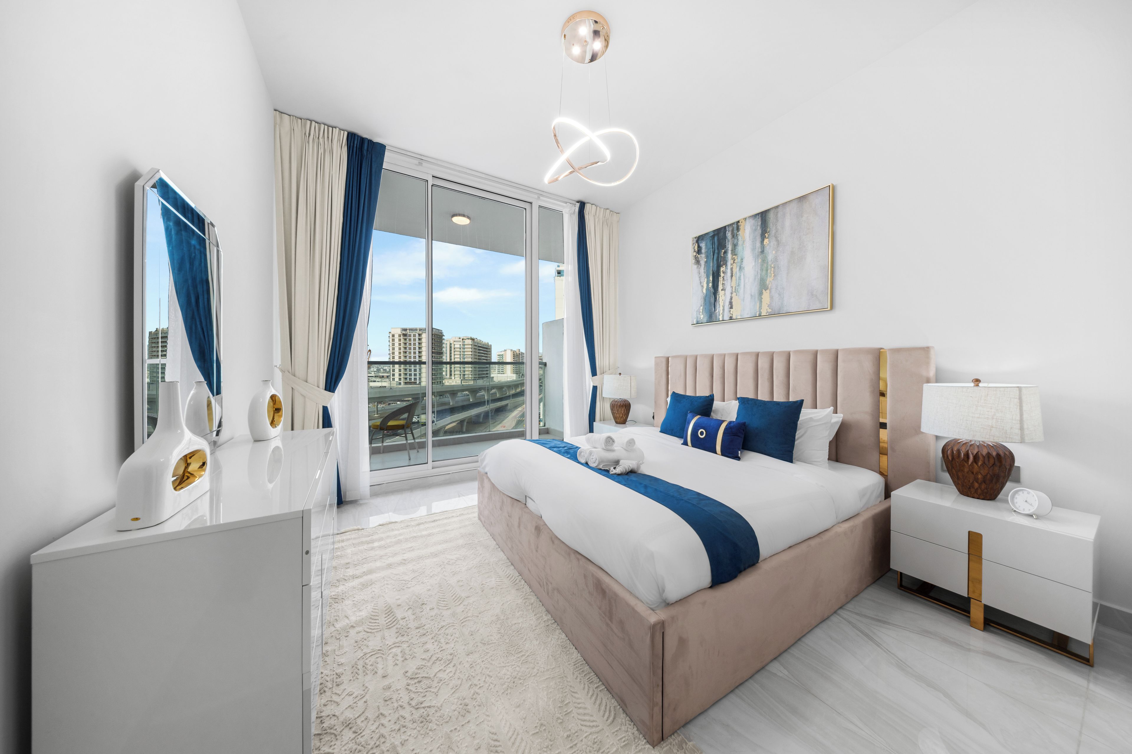 A modern hotel room with a large bed, a desk, a chair, a nightstand, a lamp, and a painting on the wall.