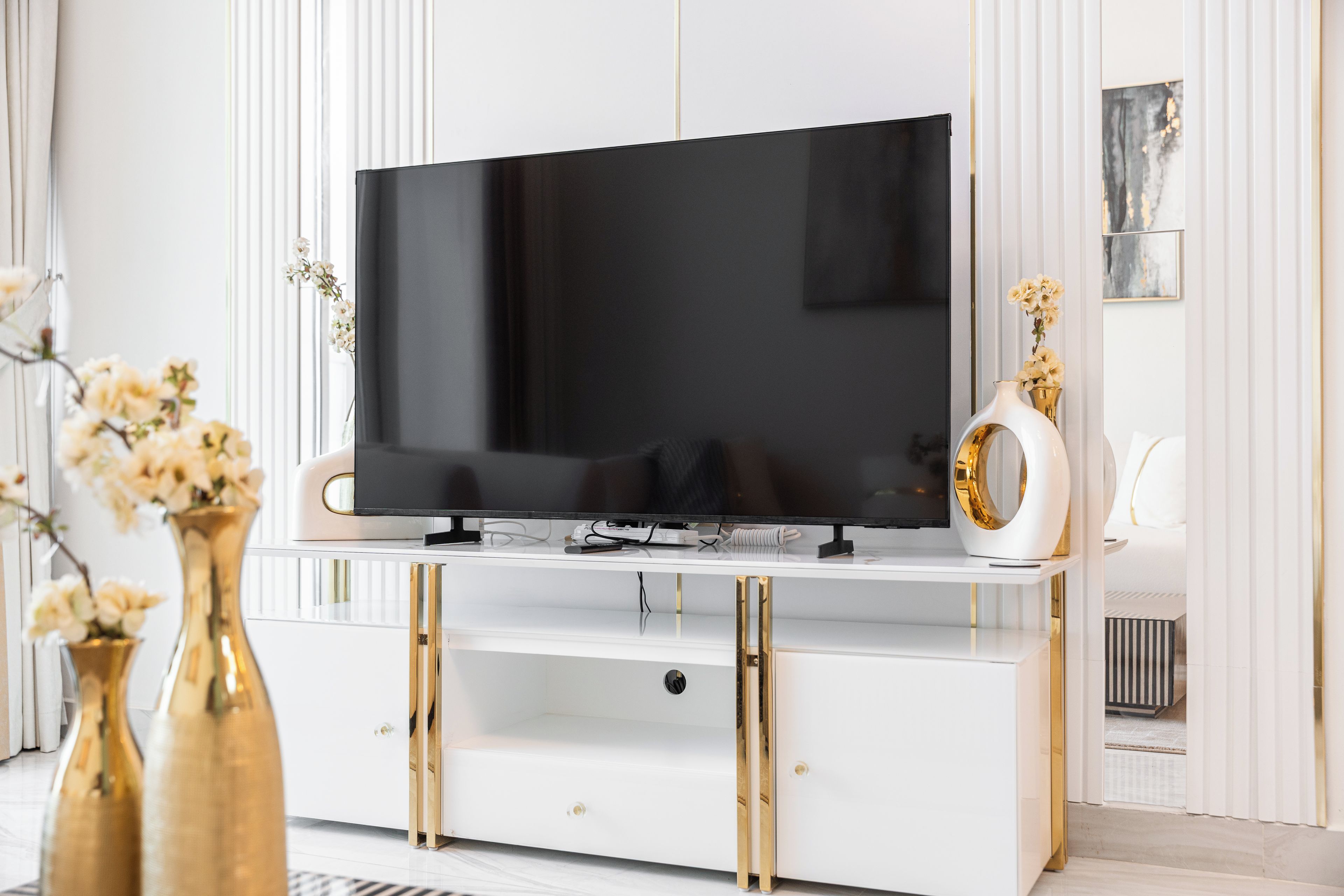 A modern living room with a flat-screen TV on a white entertainment unit with gold accents, flanked by white walls and minimal decor.