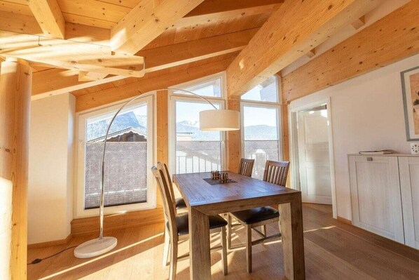 A wooden interior with a dining table, chairs, a floor lamp, and a view of mountains through large windows. - Holiday apartment with 1 bedroom (Garmisch-Partenkirchen)