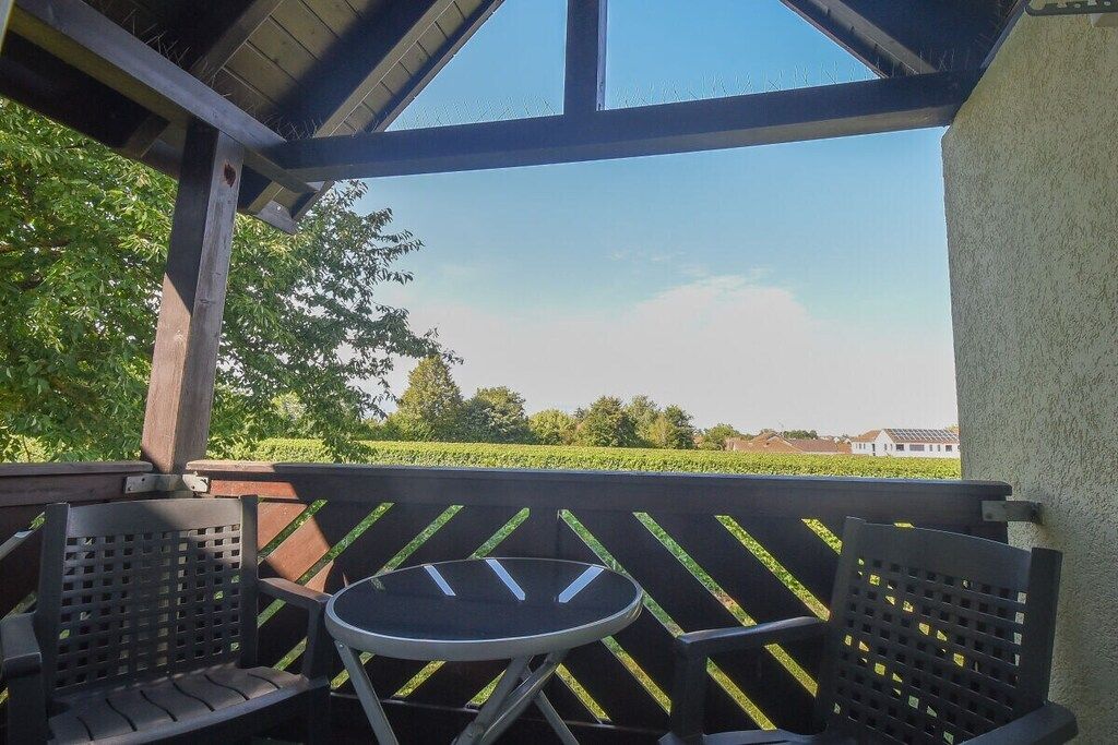 Un espace extérieur abrité, aménagé avec deux chaises et une petite table, d’où l’on peut admirer un paysage verdoyant et des bâtiments situés au loin.