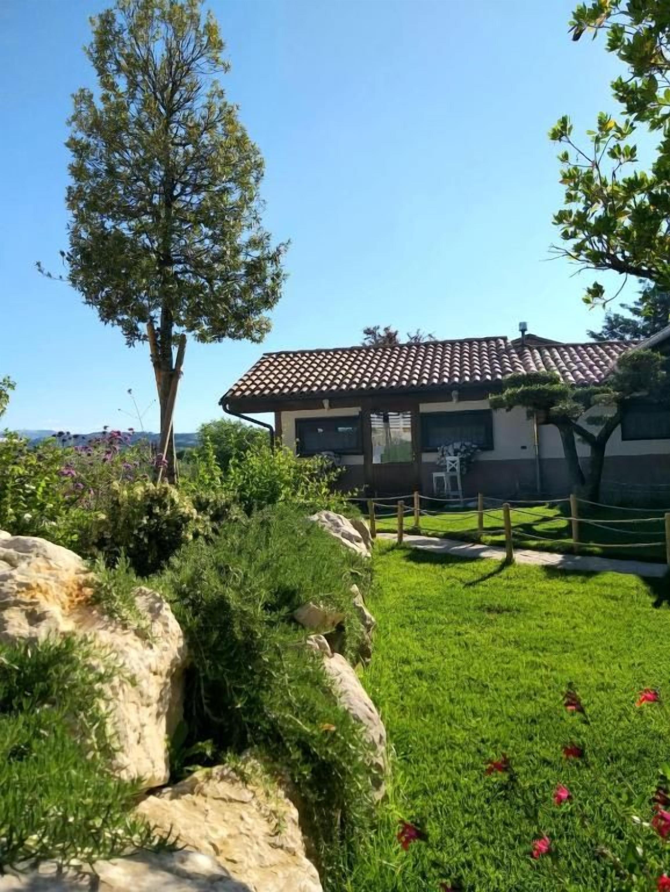 Ein Gebäude mit einem Ziegeldach, umgeben von einer gepflegten Rasenfläche mit roten Blüten, und ein großer Baum im Vordergrund.
