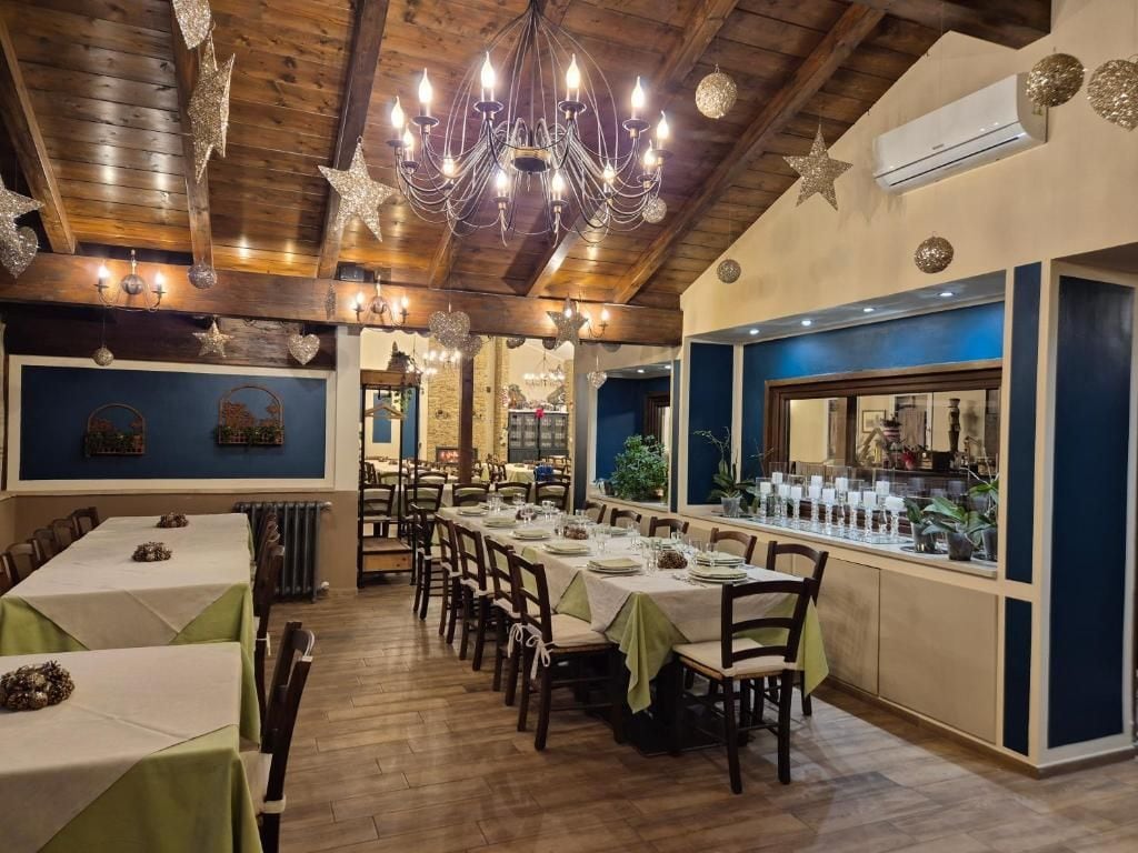 Un restaurant doté d’une longue table à manger, de nombreuses chaises et d’un lustre. On y trouve également un bar avec un comptoir et des tabourets. La pièce est ornée de poutres en bois au plafond et d’un mur en pierre à l’arrière.