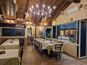 A restaurant with a long dining table, multiple chairs, and a chandelier. There is a bar area with a counter and stools. The room has wooden beams on the ceiling and a stone wall in the background.