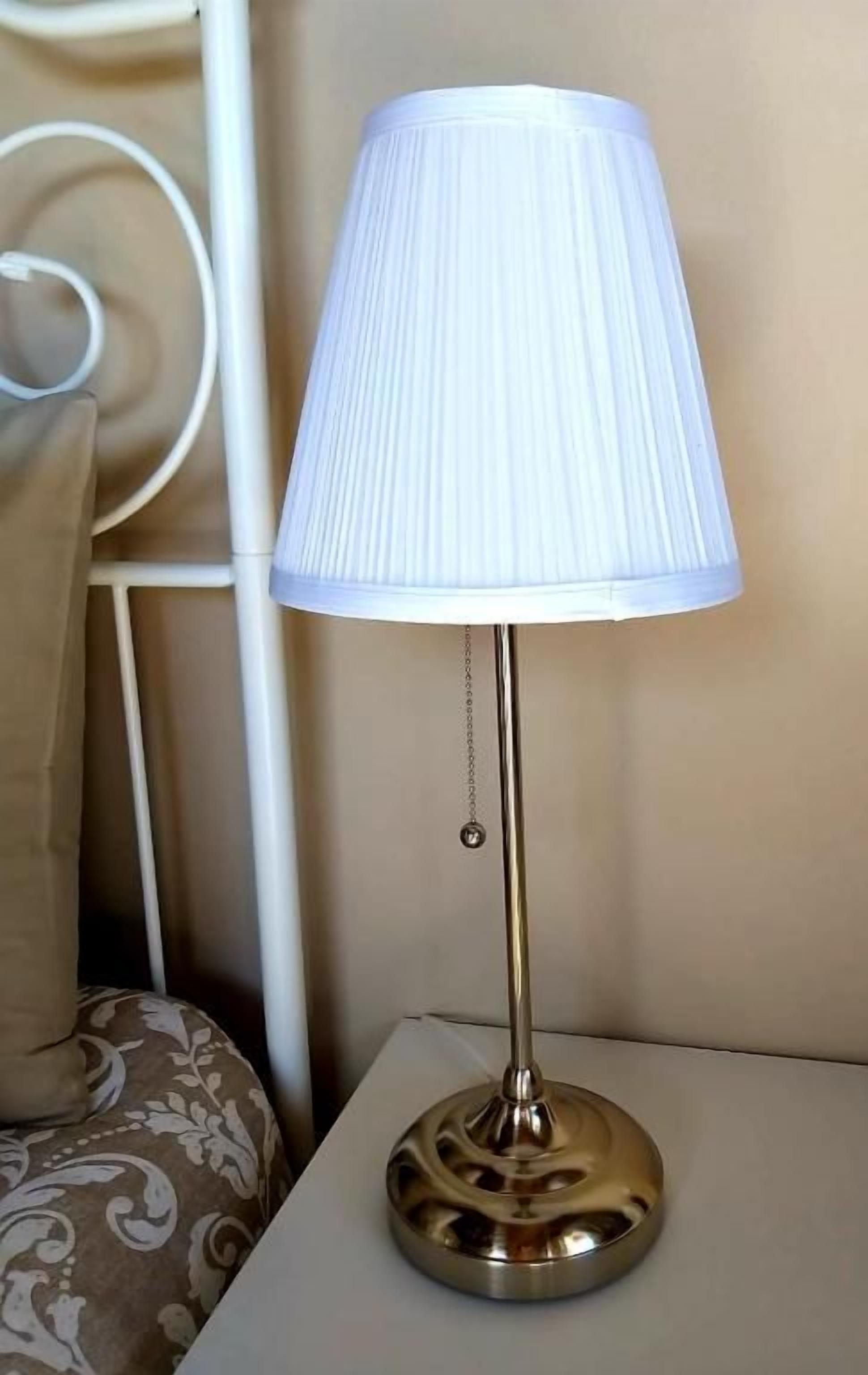 A lamp on a bedside table with a white lampshade and a metallic base.