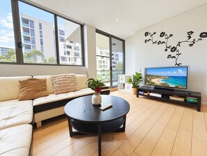 A modern living room with a sofa, coffee table, TV, and large windows. - Light Filled Zetland Retreat (Zetland)