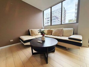 Un salon moderne avec un canapé d’angle, une table basse ronde et une grande fenêtre donnant sur des immeubles.