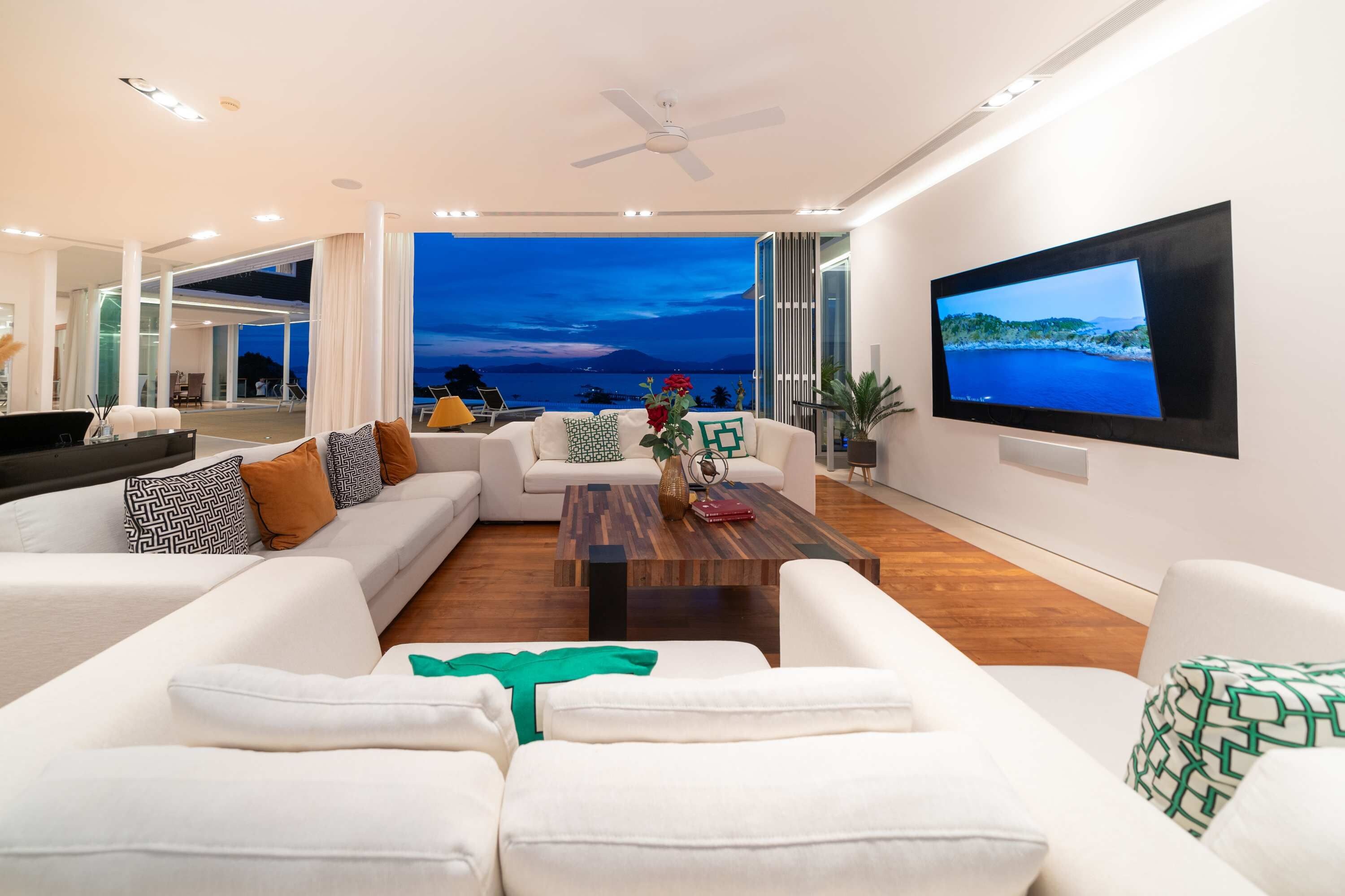 Ein modernes Wohnzimmer mit einem großen Flachbildfernseher, weißen Sofas, einem Holztisch und Blick auf ein Gewässer im Abendlicht.