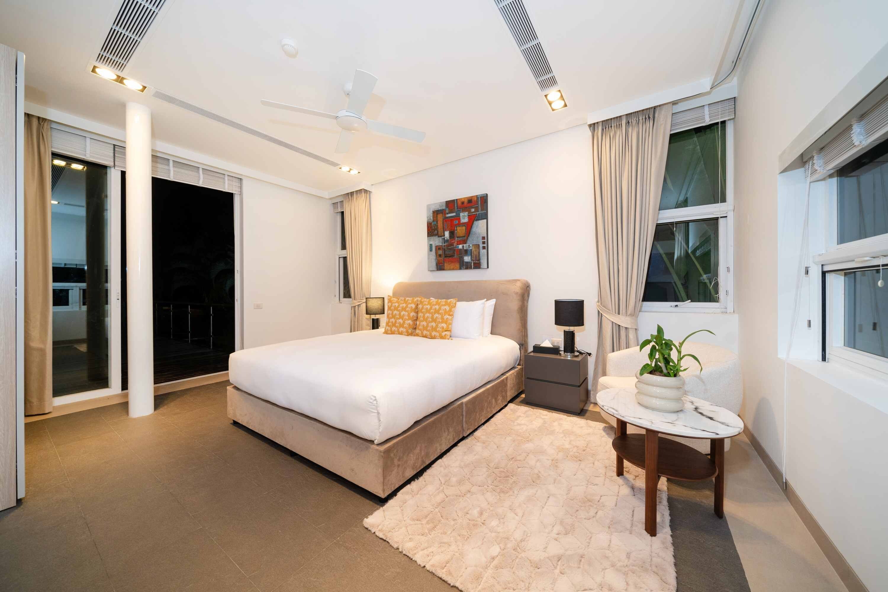 A modern bedroom with a large bed, a ceiling fan, a nightstand with a lamp, and a window with curtains.