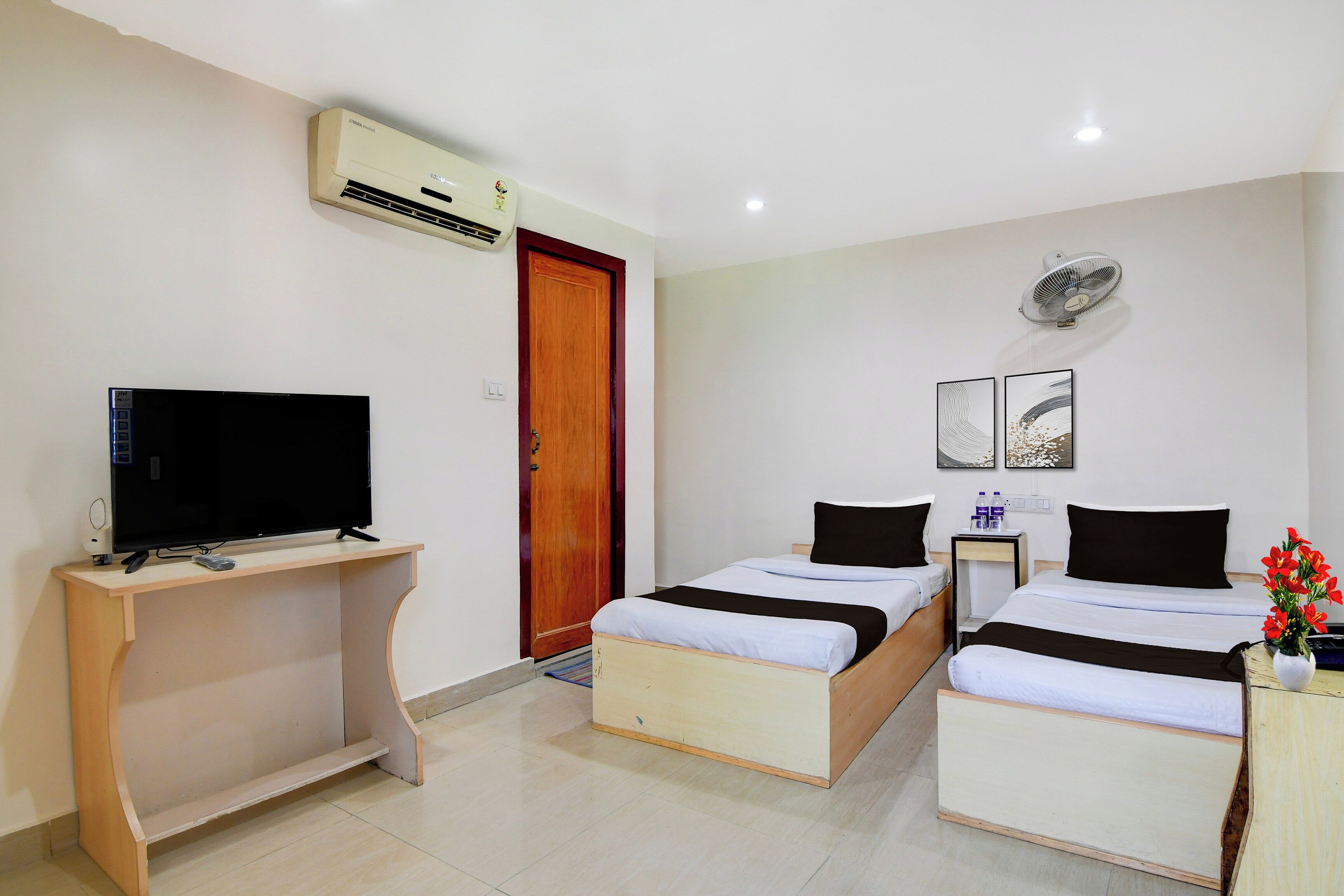 Habitación de hotel con dos camas individuales, un escritorio con televisor, aire acondicionado, ventilador de pared y una puerta.