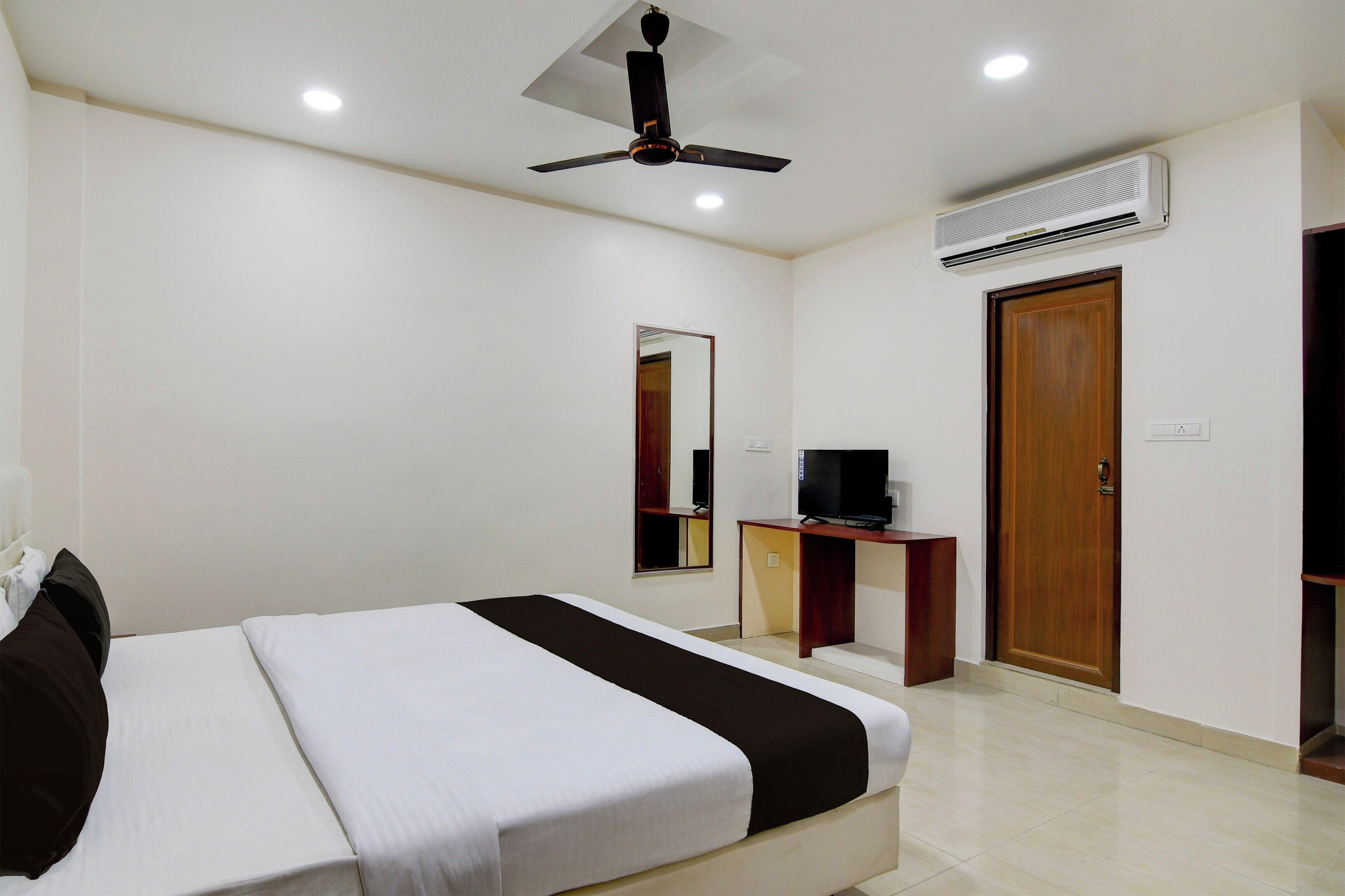 A hotel room with a bed, a television on a wooden stand, a ceiling fan, and an air conditioning unit.