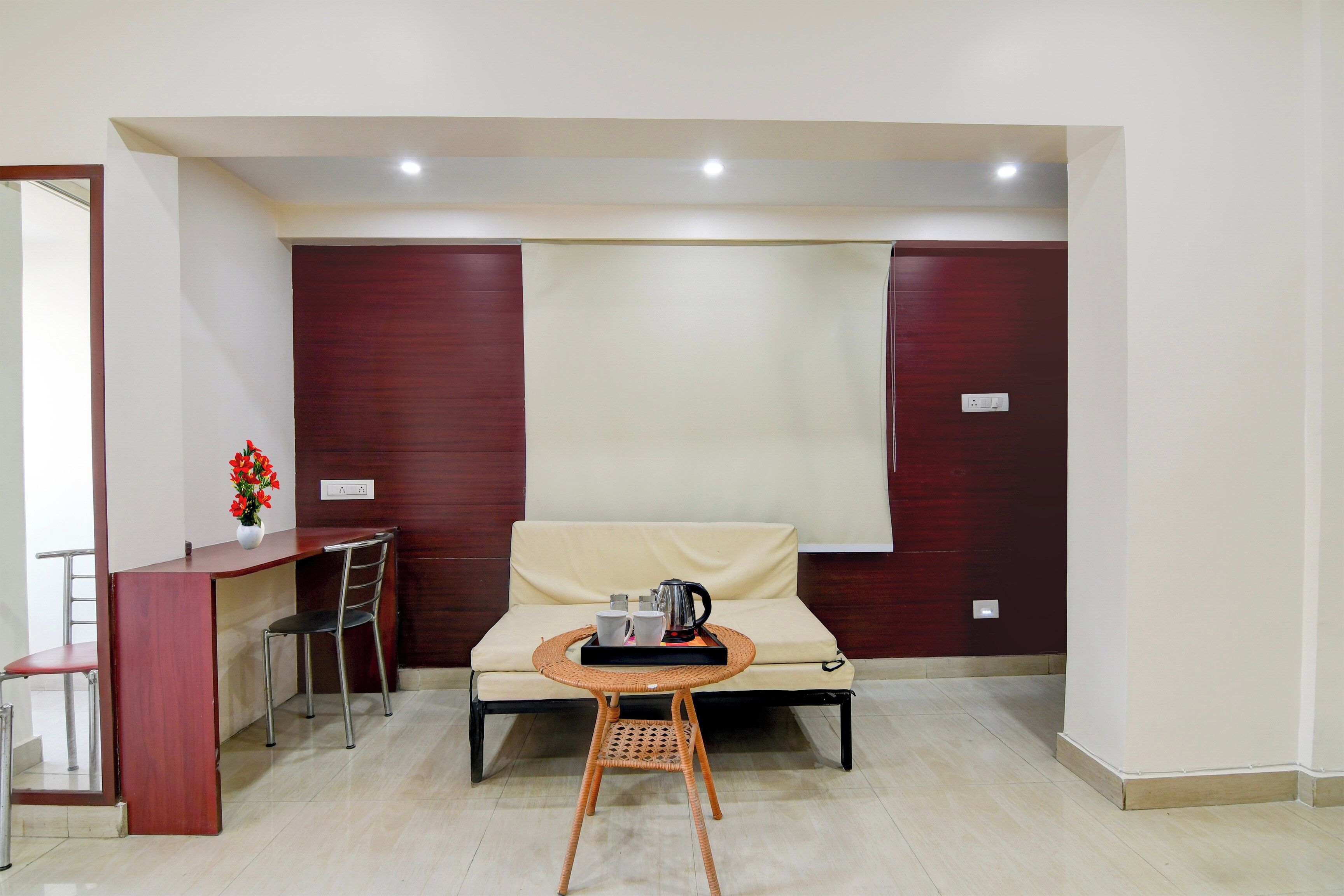A room with a sofa, a small table with a tray, a chair, a desk, and a vase with flowers.