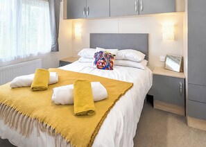 Ein ordentlich bezogenes Bett mit einer gelben Tagesdecke und zwei Handtüchern, ein Nachttisch mit einem gerahmten Bild und ein Fenster mit Vorhängen.