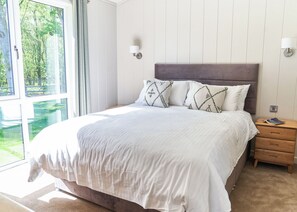 A bedroom with a bed, two bedside tables, a window with curtains, and a view of greenery outside. - Autograph Retreat Hot Tub 2 bed Luxury Lodge slps4 (Landford, Nr Salisbury)