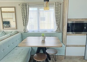 A modern kitchen with a built-in oven, a dining area with a wooden table and two stools, and a large window with curtains. - Gold 8 berth Pet Friendly (Chapel St Leonards)