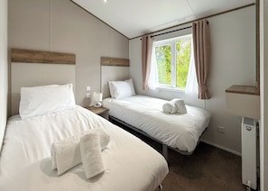 A hotel room with two beds, a window with curtains, a bedside table with a lamp, and a small heater. - Woodland 3 bed Lodge - Hazel (Notter Bridge, Saltash)
