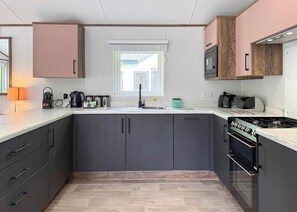 A modern kitchen with grey cabinets, a built-in oven, and a window above the sink. - Woodland 3 bed Lodge - Hazel (Notter Bridge, Saltash)