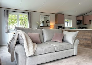 A modern living room with a grey sofa, a kitchen area with wooden cabinets, and a large window with curtains. - Woodland 3 bed Lodge - Hazel (Notter Bridge, Saltash)