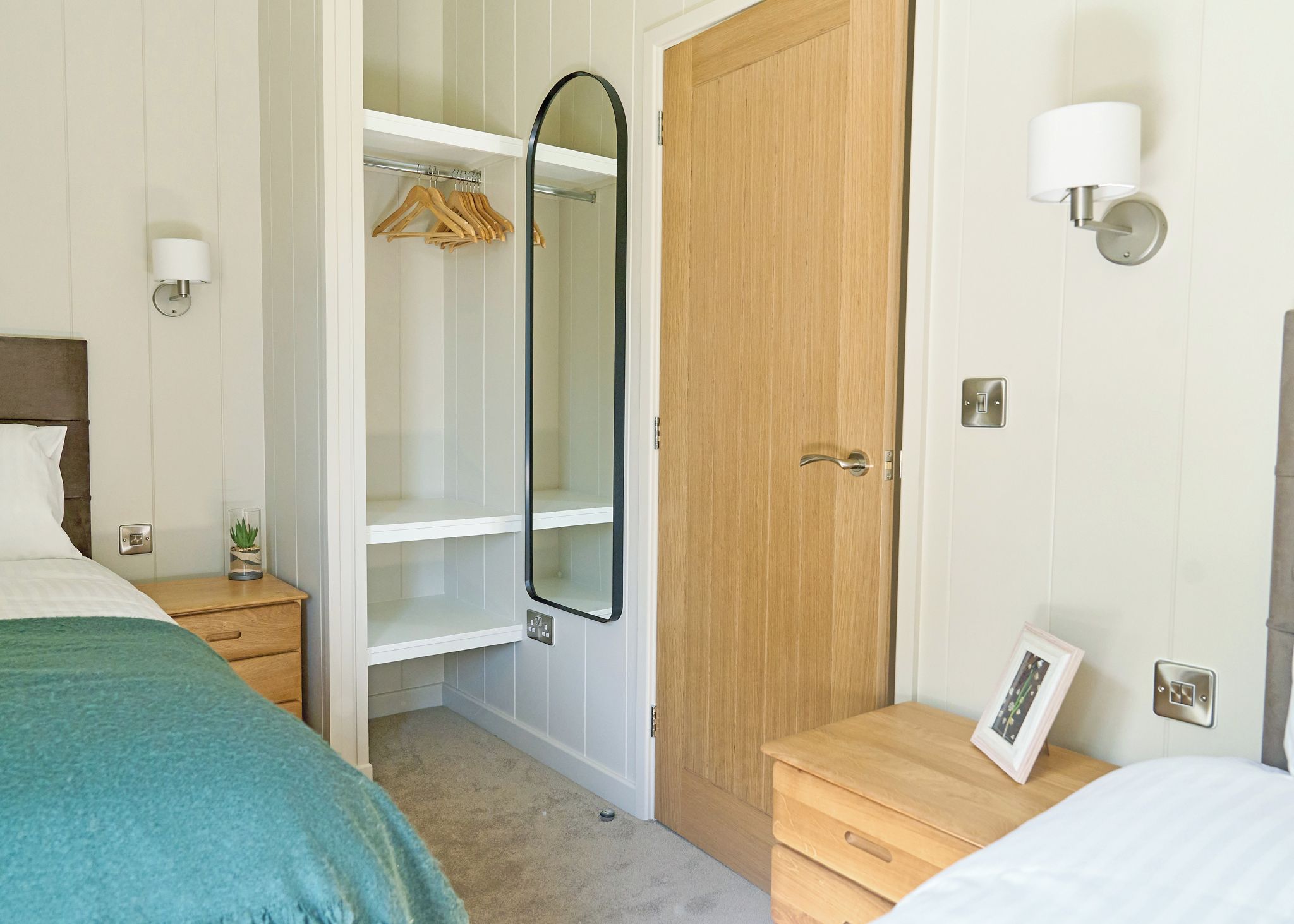 Een slaapkamer met een bed, een nachtkastje, een wandlamp, een houten garderobe en een spiegel.