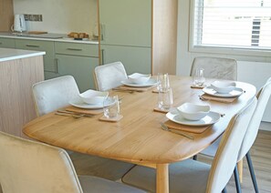 Mesa de jantar posta para seis lugares, com pratos, tigelas e talheres brancos, e uma janela com persianas ao fundo.