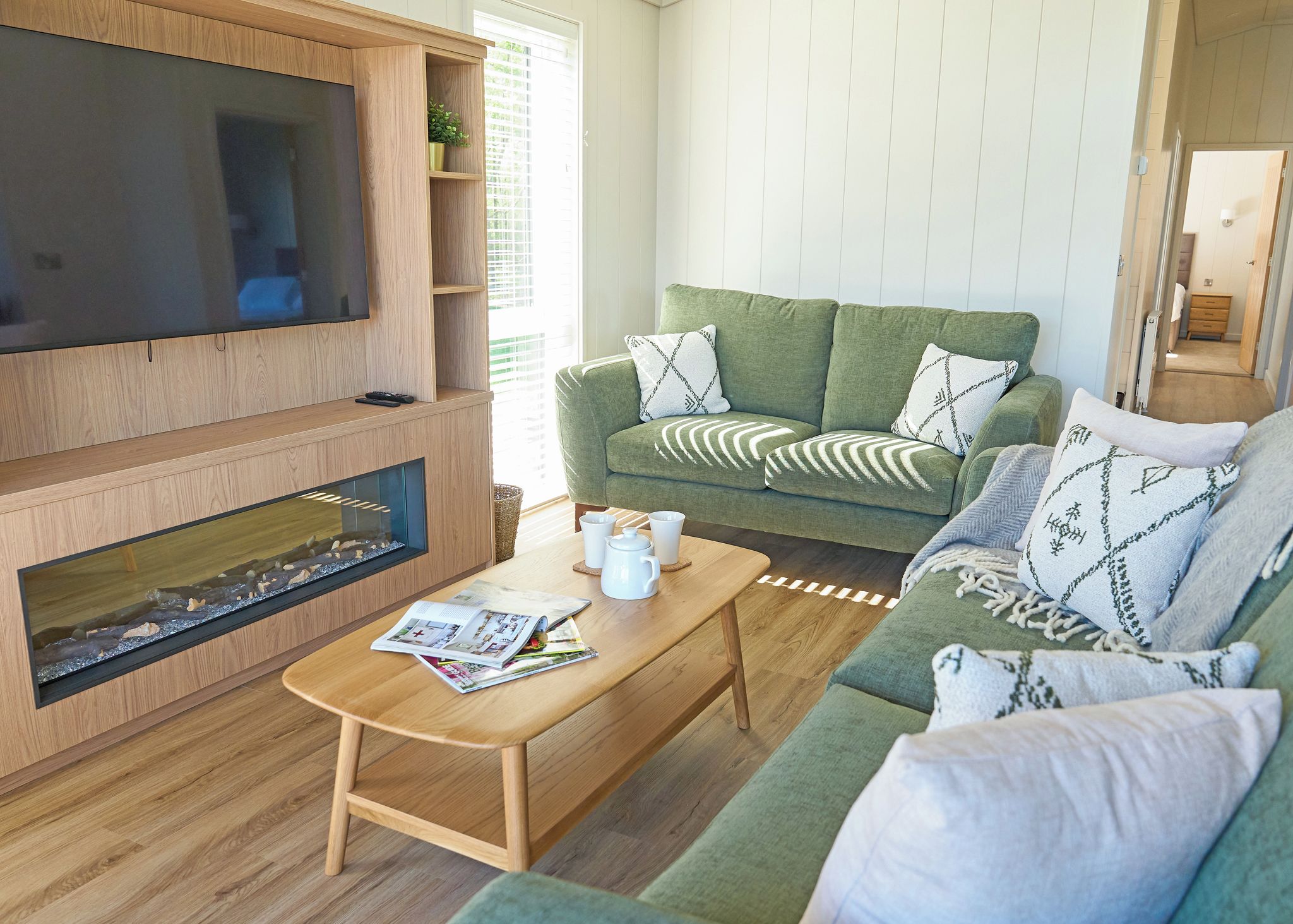A living room with a flat-screen TV, a fireplace, a green sofa with patterned cushions, and a coffee table with magazines and a teapot.