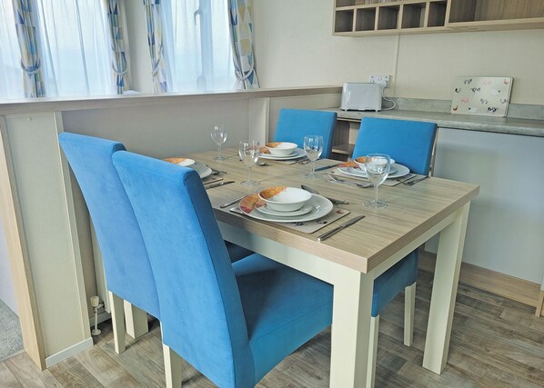 Comedor con una mesa dispuesta para cuatro, sillas azules y una zona de cocina con decoración mural con motivos de frutas.