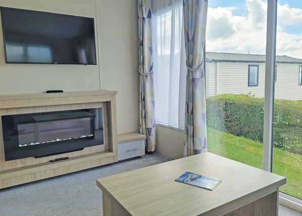 Een kamer met een wandgemonteerde flatscreen-tv, een ingebouwde open haard, een tafeltje met een tijdschrift en een raam met gordijnen.