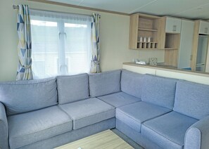 A modern living room with a grey sectional sofa, a wooden cabinet with glassware, and a window with patterned curtains. - 1 bedroom accommodation in Dorchester (Dorchester)