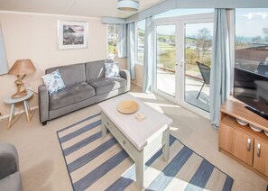 A modern living room with a grey sofa, a small table, a TV, and a view of the outdoors through large windows. - 2 bedroom accommodation in Budleigh Salterton (Budleigh Salterton)